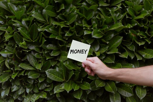 A hand holding a note with 'MONEY' written on it in front of lush green leaves.