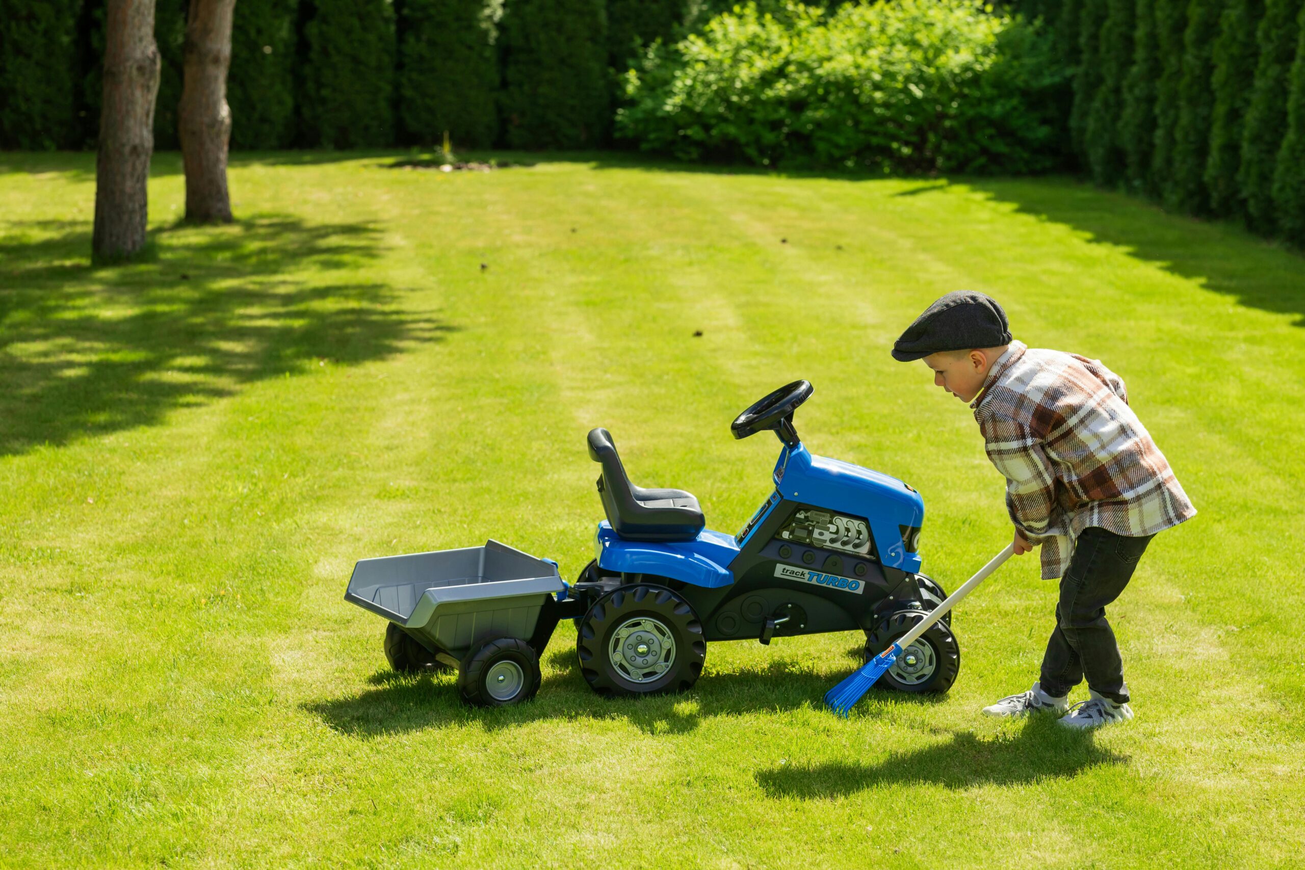 A young boy with a cap plays with a blue toy tractor and broom on a sunny lawn.